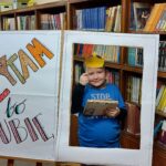Chłopiec z książką pozuje przy napisie "Czytam bo lubię". Nad głową trzyma papierową koronę