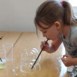 Uczennica tworzy na ławce szkolnej bańki mydlane za pomocą słomki. Udaje jej zamknąć bańkę w bańce.